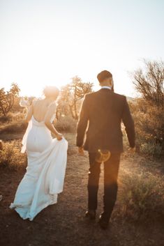 a bride and groom walking through the desert at sunset in their wedding attire, holding hands