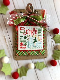 a cross stitch christmas ornament hanging on a wooden table with holly and bells