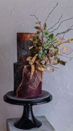 a three tiered cake decorated with leaves and flowers on a black stand against a white wall