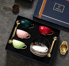 there are many cups and saucers in the box on the table with gold spoons