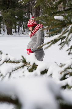 a person in the snow with a hat and scarf around their neck, standing next to a pine tree