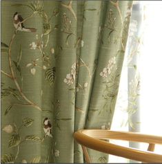 a chair sitting next to a window covered in green floral curtain panels with birds on them