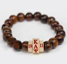 This exquisite bracelet features rich brown beads, polished to a lustrous sheen, crafted from natural wood. The centerpiece is a trio of unique golden beads, each engraved with a special character: the Greek letters Kappa Alpha Psi flanked by the Founding Year ‘19’ and ‘11’. This design combines elegance with a hint of personal significance, making it a perfect accessory for those who appreciate meaningful jewelry. Kappa Alpha Psi, Alpha Phi Alpha, Golden Beads, Omega Psi Phi, Wood Bead Bracelet, Promo Items, Greek Letters, Silicone Bracelets, Meaningful Jewelry