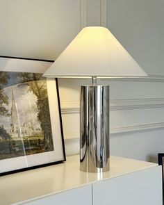 a lamp sitting on top of a white dresser next to a painting and framed pictures