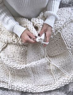 a woman is holding a white object in her hands while sitting on a chair with a knitted blanket around her