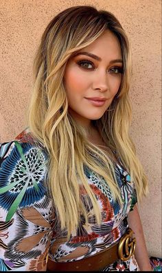 a woman with long blonde hair wearing a colorful shirt and brown belted dress standing in front of a wall