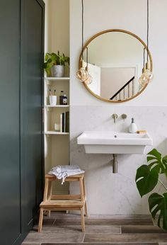 a white sink sitting under a round mirror next to a wooden stool and book shelf