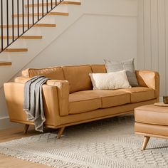 a tan leather couch sitting in front of a stair case next to a chair and ottoman