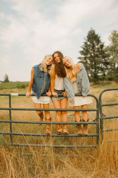 three young women standing on top of a metal fence in a field with tall grass