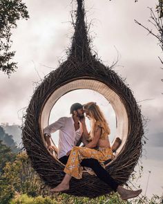 a man and woman sitting in a bird nest on top of a hill with trees