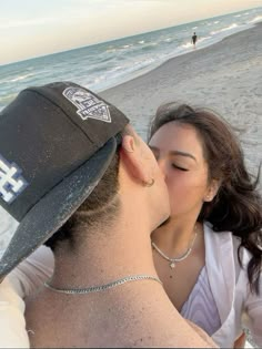 a man kissing a woman on the beach while wearing a baseball cap and diamond necklace