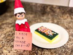 an elf holding a sign next to a piece of cake with sprinkles on it