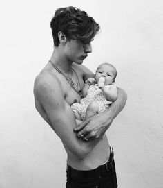 a young man holding a baby in his arms and looking at the camera while standing against a white wall