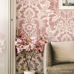 pink flowers are in a white vase next to a gray chair and wallpapered with an ornate pattern