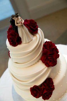 a white wedding cake with red roses on top and a bride and groom figurine