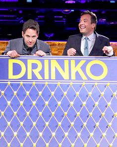 two men standing behind a sign that says drinko on the front, and one man in a suit is smiling