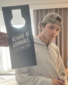 a young man in a gray hoodie is holding up a cardboard sign that says beware of adorable