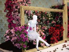 a white mannequin sitting in front of a mirror with pink and red flowers