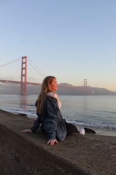 a woman sitting on the edge of a concrete wall next to water and a bridge