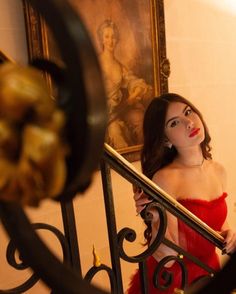 a woman in a red dress is standing on a stair case and looking into the mirror