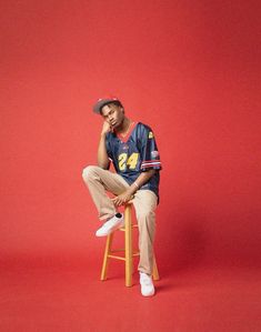 a man sitting on top of a wooden stool in front of a red wall wearing a hat