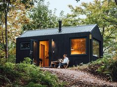 a man sitting on a chair in front of a small black cabin surrounded by trees