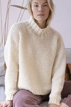 a woman sitting on top of a bed wearing a white sweater and purple pants with her hands in her pockets