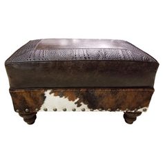 a brown and white cowhide ottoman with metal studs on the bottom, sitting on top of a wooden base