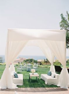 an outdoor wedding setup with white drapes and couches on the grass, overlooking the ocean