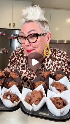 a woman with white hair and glasses sitting in front of trays of cupcakes