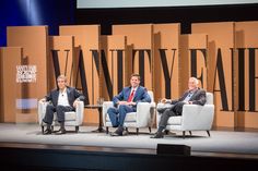 three men sitting on chairs in front of a stage with the words vanity fair written on it