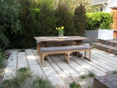 a wooden table and bench in a small backyard area with stone flooring, grass and flowers