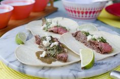 three steak tacos on a marble plate with lime wedges next to the plates