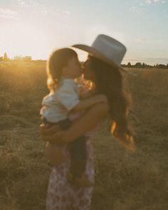 a woman holding a baby wearing a cowboy hat in a field with the sun behind her