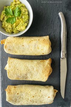 three burritos on a slate board next to a bowl of guacamole and a knife