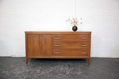 a wooden dresser with two drawers and a vase on it's top, against a white brick wall