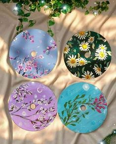 four circular discs with flowers painted on them sitting next to a green plant and white background