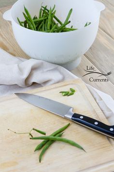 green beans are sitting on a cutting board next to a knife and bowl full of them