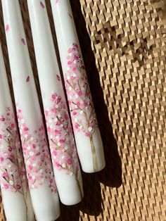 four white candles with pink flowers painted on them sitting on a woven tableclothed surface