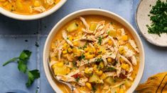 two bowls of chicken and corn soup on a blue table with spoons, parsley