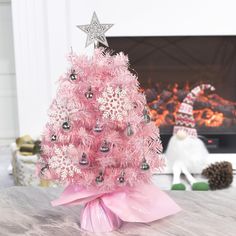 a pink christmas tree decorated with silver ornaments and snowflakes, sits on a table in front of a fireplace