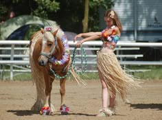 a woman in a hula skirt standing next to a brown horse on a dirt field