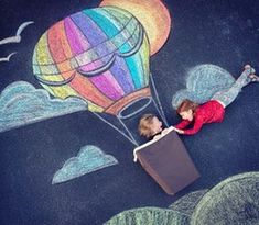 two children are playing with chalk drawings on the ground while an air balloon is in the sky