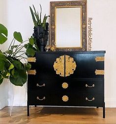 a black and gold dresser with mirror on top next to potted plant in front of it