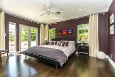 a large bedroom with hardwood floors and purple walls, along with two windows that look out onto the backyard