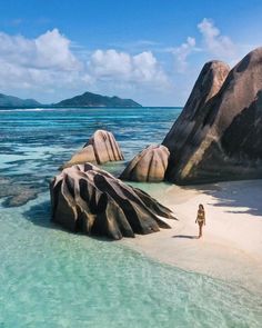 a woman standing on the beach next to large rocks in the ocean with clear blue water