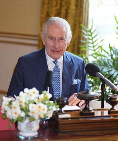 an older man wearing a suit and tie sitting at a desk with microphones in front of him