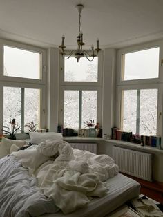 an unmade bed in front of three windows with books on the floor and a chandelier