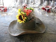 a hat with sunflowers and other flowers in it sits on a table surrounded by wine glasses