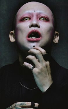 a man with red makeup and piercings holding his hand up to his face while looking at the camera